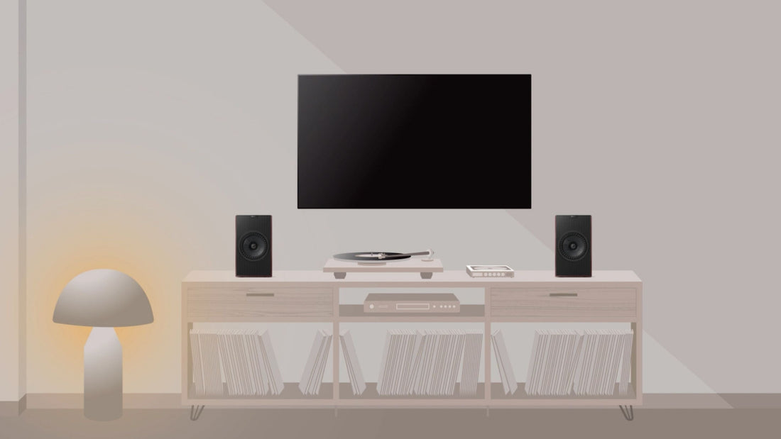 A minimalist living room setup featuring a black flat-screen TV mounted on a light beige wall above a white media console. The console holds a turntable, a pair of Coda W speakers, and a stack of books or records.