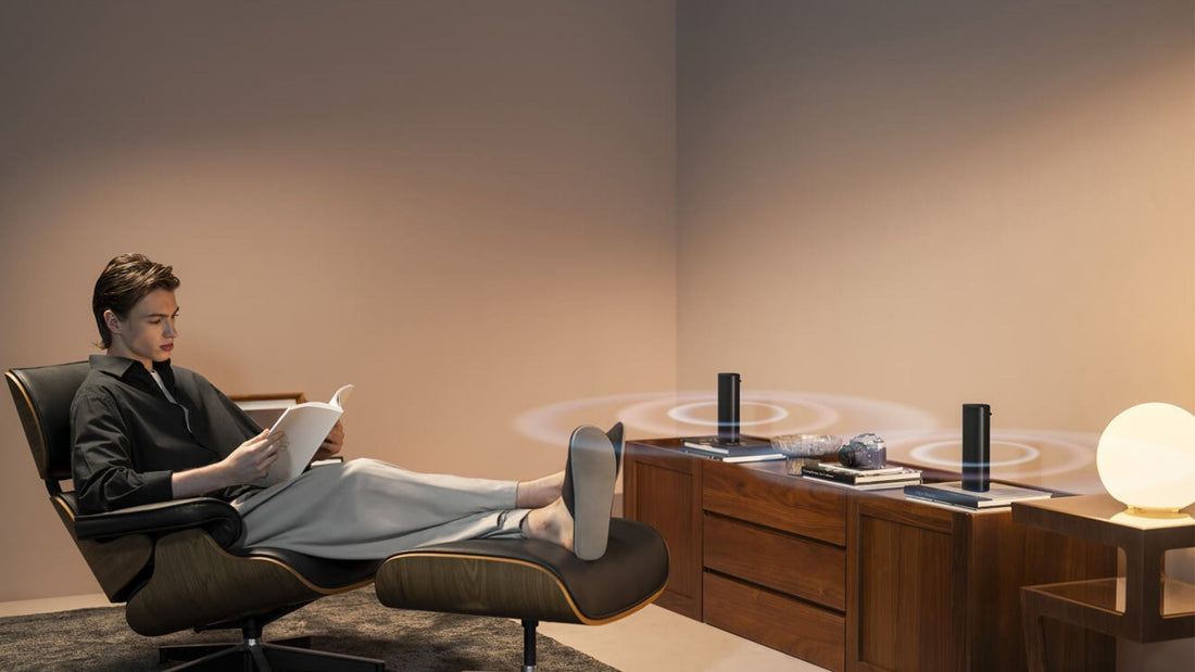 A man comfortably sitting in a chair, focused on a book in his hands, listening to KEF Muo portable speakers with dual stereo mode 